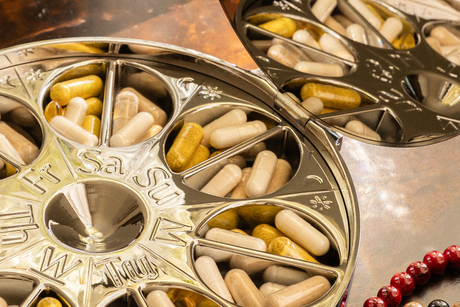 A round silver compartment pill organizer filled with beige and yellow capsules, styled on a dark wooden table with a matching silver lid, a glass vase, and a leather journal nearby.