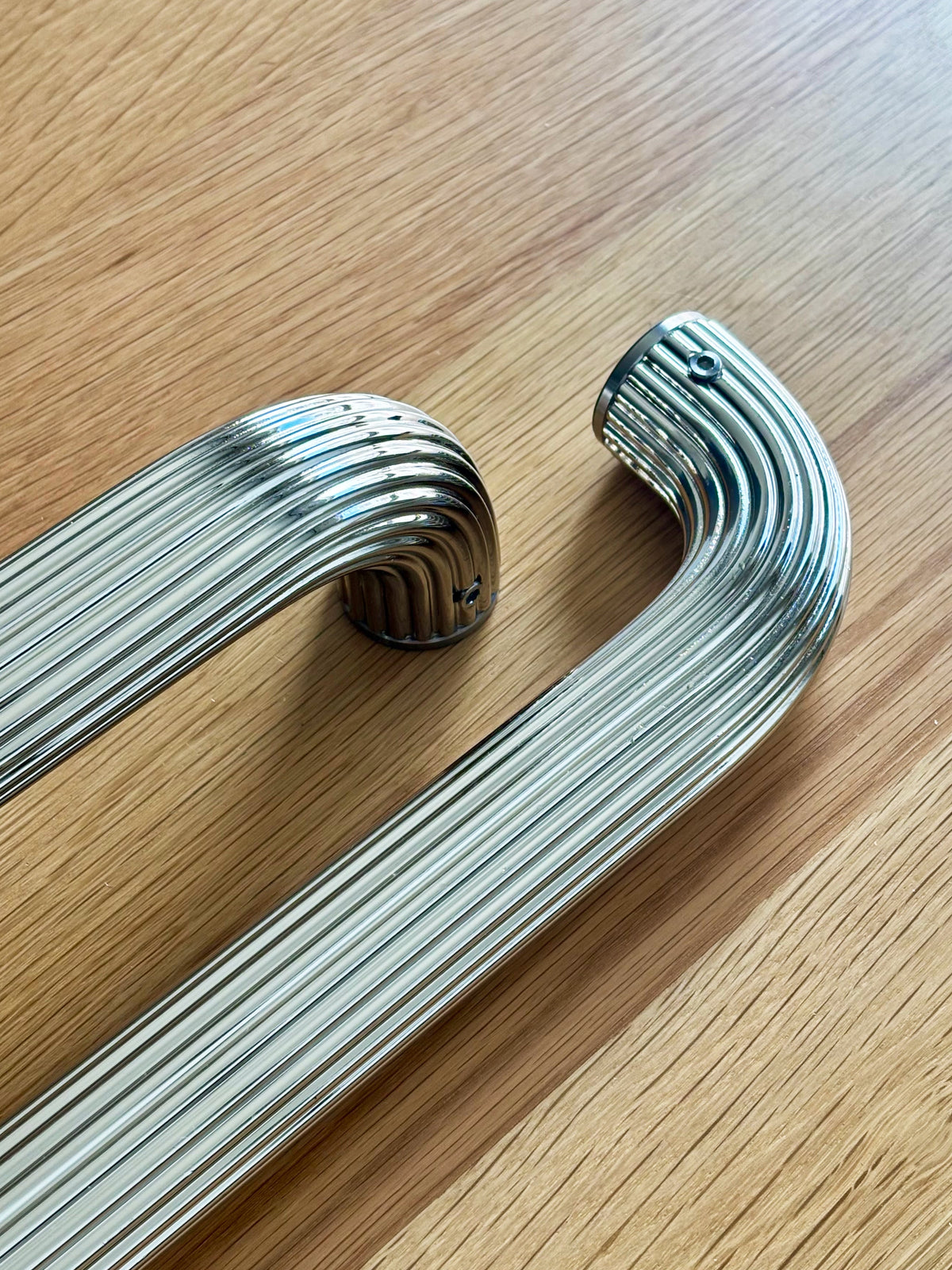 Two polished chrome grab bars with a ribbed, curved design, displayed on a light wood grain surface optimized for universal design.