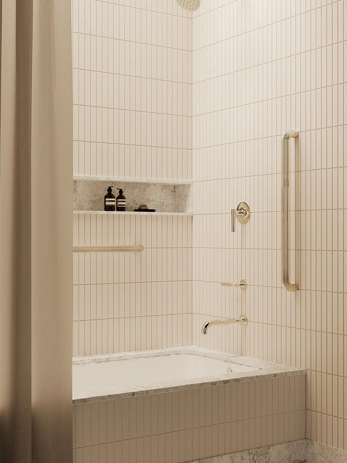 A modern bathroom corner with cream vertical subway tiles, a white bathtub with a marble-look surround, and warm polished nickel fixtures including the grab bar designed by REMSEN.